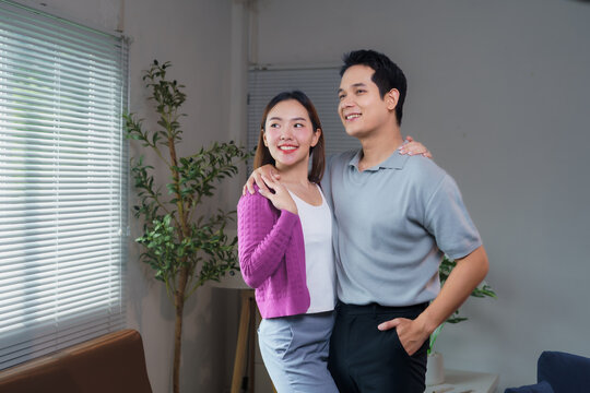 Young Asian couple embracing and gazing out the window, savoring the joy of their new home together in a cozy living room