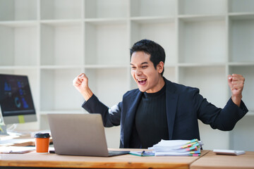 Young asian manager raising his arms and screaming while looking at his laptop, celebrating a...