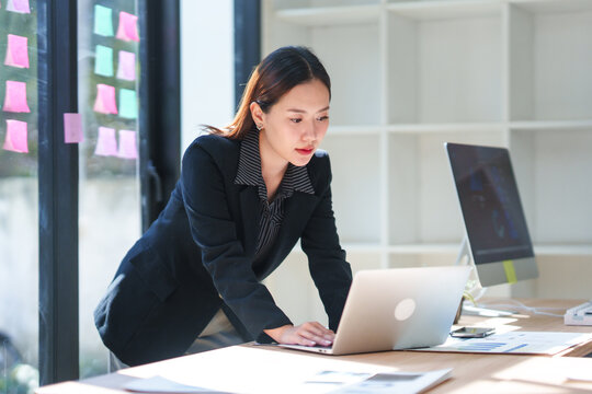 Asian businesswoman standing and working on laptop in modern office with sticky notes on window