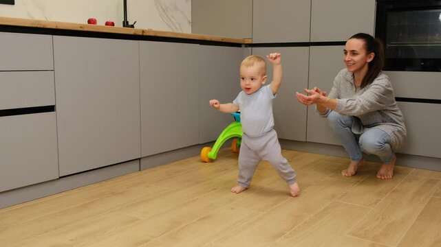 The child learns to take the first steps without mother's help. little boy he takes a few steps and falls down confused. Mom helps the baby learn to walk in kitchen mother support young toddler son