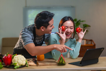 Young cheerful asian couple wearing aprons having fun and preparing healthy meal together in kitchen, using online recipe on tablet