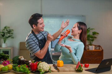 Young asian couple having fun singing with carrot like microphone while preparing healthy meal in modern kitchen