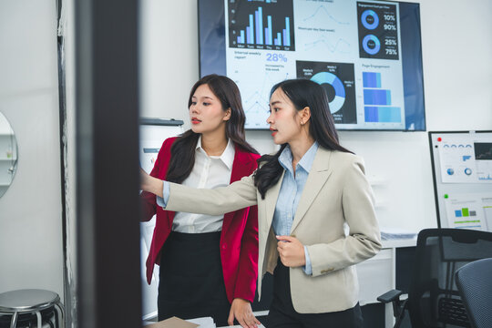 Two asian businesswomen discussing marketing strategy while analyzing financial charts and graphs displayed on a whiteboard and interactive screen during a meeting in a modern office