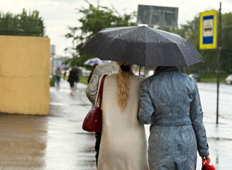 Women walk along the sidewalk under a black umbrella. Rainy weather in a European city. A women...