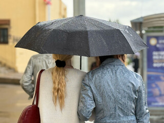Women walk along the sidewalk under a black umbrella. Rainy weather in a European city. A women...