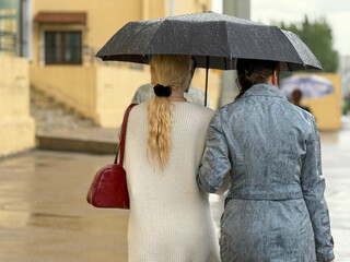 Women walk along the sidewalk under a black umbrella. Rainy weather in a European city. A women...