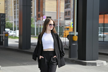 Woman walking in stylish outfit holding jacket on shoulder in urban environment on sunny day.