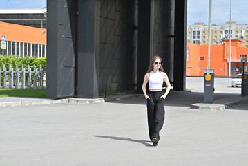 Woman walking in stylish outfit holding jacket on shoulder in urban environment on sunny day.