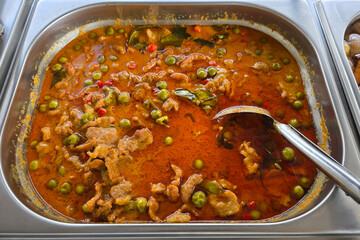 Thai-style pork curry served in a stainless steel tray, featuring rich red curry sauce with sliced pork, pea eggplants, chili, and kaffir lime leaves.