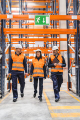 Logistics workers wearing safety vests and helmets are walking through a warehouse, using handheld devices to check inventory and manage goods
