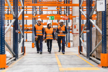 Warehouse workers wearing safety vests and helmets are walking through a large distribution center, discussing logistics and inventory management