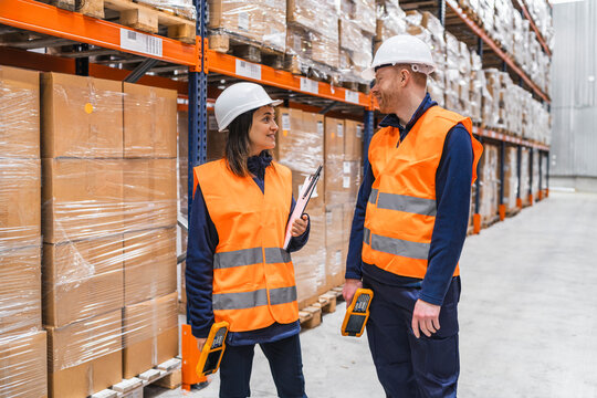 Two warehouse workers wearing safety vests and hard hats are discussing logistics while holding handheld devices, surrounded by shelves full of packaged goods