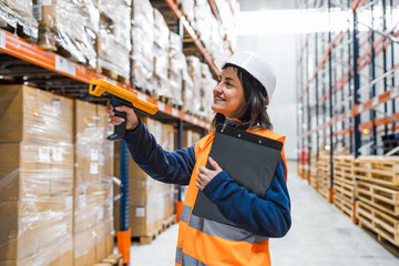Smiling female warehouse worker wearing safety helmet and vest, holding clipboard and using barcode scanner for inventory management in large distribution center
