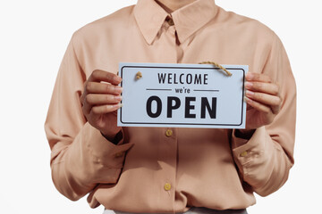 A confident young Asian woman in a light brown shirt stands against a white background, holding a white &ldquo;Welcome, We Are Open&rdquo; sign, warmly inviting customers to enter with a friendly smile.