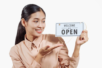 A confident young Asian woman in a light brown shirt stands against a white background, holding a white &ldquo;Welcome, We Are Open&rdquo; sign, warmly inviting customers to enter with a friendly smile.
