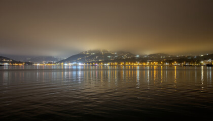 Obraz premium Calm water reflects city lights with foggy hills creating peaceful night scene