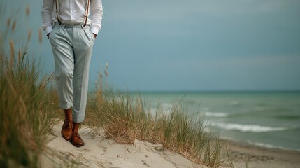 A man in light blue pants and suspenders walking on a sandy beach near the ocean water and tall grass