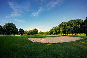 Wide-angle view of a golf green with flagstick, trees, and bunker under a vibrant blue sky. Perfect...