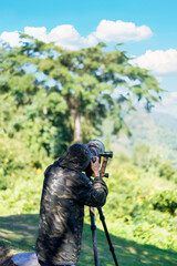 Man, photographer is wearing camouflage jacket taking photos with Dslr camera and telephoto lens in green forest at Khao Yai's 30th km Viewpoint in the Khao Yai National Park, Thailand.Vertical.