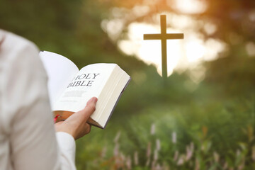 Christian woman holding bible and reading word of God in forest with brightly lit church cross, faith and belief background
