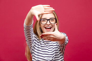 One Happy Winsome Excited Energetic Girl Sitting on Floor With Glasses And Making Frame With Hands As Celebration Or Winning Isolated on Crimson Background.