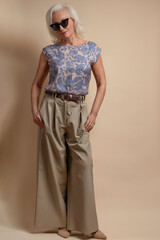 Full Length Portrait of Tranquil Senior Caucasian Blond Woman Posing in Sunglasses in Casual Floral Blouse And Beige Pants In Studio Over Beige Background With Hands in Pockets