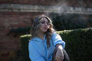 Young, pretty, blonde woman, wearing a shirt, exhaling tobacco smoke through her mouth, sitting in...