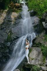 Woman practicing yoga near a majestic waterfall surrounded by lush green forest and rocky cliffs under natural daylight in an outdoor serene setting.