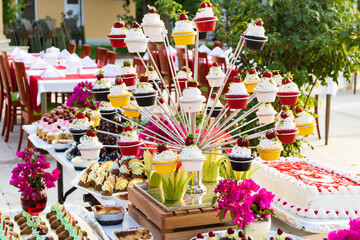 A vibrant arrangement of colorful cupcakes on a tiered display surrounded by various desserts including cakes, pastries, and fresh fruits. Bright pink flowers and elegant table setups enhance the