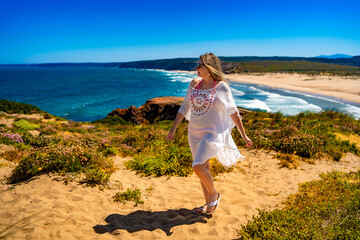 Beautiful middle aged woman walking in flowing dress at viewpoint on sandy beach Bordeira Praia in...