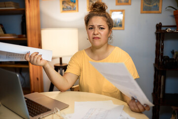 Horizontal image of worried woman in yellow t-shirt trying to cope with documents working online from home, sitting at table in living-room using laptop, having troubles with work task