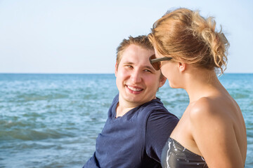 A smiling young guy and a girl are talking on the beach against the background of the blue sea and blue sky on a sunny summer day