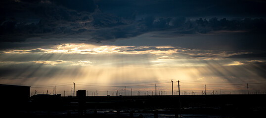 The photo shows industrial buildings and power lines against the sunset. Golden rays of light break through the clouds, creating a dramatic and atmospheric scene.