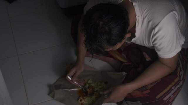 A poor man wearing a white shirt is eating hungrily from food placed on the floor over a piece of paper. His intense focus and quick movements reflect deep hunger. The scene captures a raw and emotion