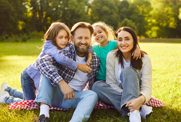 Fototapeta premium Happy family sitting together on plaid in nature, enjoying picnic time in park, smiling gladfully, having nice time together during summer, taking memorable photo. People, rest, vacation 