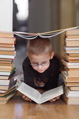 A young boy with glasses lies on the floor surrounded by stacks of books, reading with curiosity...