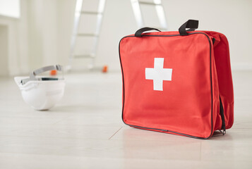 Closeup of red bag with first aid kit standing in empty building site with safety helmet and construction ladder on floor. Emergency, health insurance and job accident at work concept.