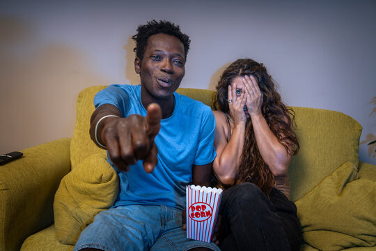 Couple watching a scary movie at home: woman hides her face in fear, man points at the screen.