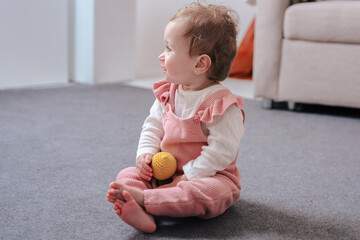 Portrait Adorable baby girl sitting on the carpet, barefoot and holding a yellow toy. Bright and...