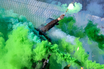 Person igniting a bright green flare with thick smoke during an outdoor event behind a metal fence