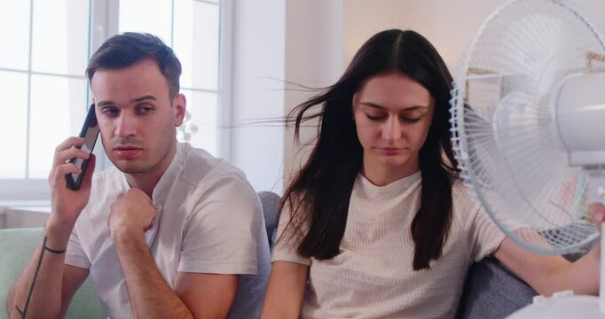Family couple having problem with air conditioning during very hot extreme summer weather heatwave. Exhausted sweaty people call repair service on mobile phone while trying to refresh by electric fan