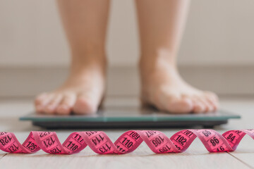 A pair of feet stands on a digital scale. A pink measuring tape is coiled in front of the scale....