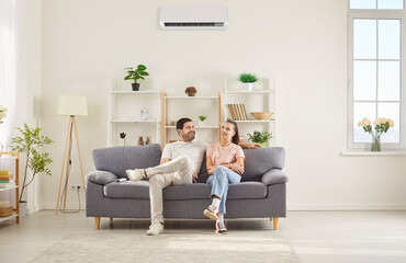 Happy couple sitting on comfortable gray sofa in living room, relaxing together under air conditioner. Smiling young man and woman resting on couch, talking and enjoying fresh cool air at home.