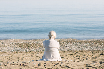 An elderly woman sits on the sand facing the calm sea, wrapped in a light cardigan. The peaceful...