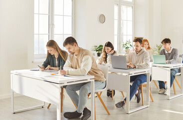 Group of young confident students sitting at the desk and writing test during a college or university lesson in classroom. People studying on project. Education and knowledge concept.