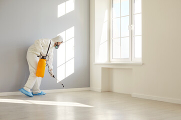 Professional exterminator in white workwear spraying insecticide in modern empty room, disinfecting...