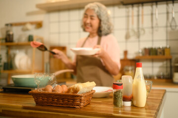 Close-up view of everyday kitchen ingredients including sauces, spices, eggs, and bread on a wooden counter, with an elderly woman happily cooking in the background