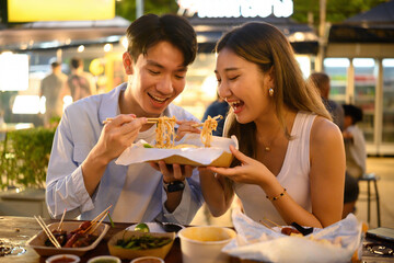A smiling couple enjoys sharing a box of noodles under vibrant night market lights. A lively and...