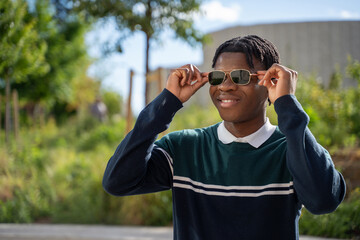 Smiling african american student putting on sunglasses in a university campus park, enjoying a sunny day