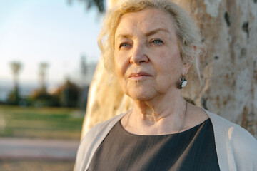 Elderly woman stands outside by a tree, looking ahead with a calm, thoughtful expression. Natural light and outdoor setting convey themes of confidence, wisdom, and graceful aging.
